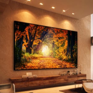 May include: A large rectangular wall art print featuring a forest path covered in fallen orange and yellow autumn leaves, framed in black, mounted on a textured beige wall above a long wooden media console with stacks of books.