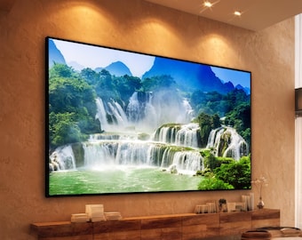 Op de afbeelding: Een grote rechthoekige wandkunstprint met een levendig landschap van een waterval met meerdere niveaus die over rotsachtige kliffen in een groene poel stroomt, omringd door weelderige groene bomen en verre blauwe bergen.