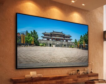 Op de afbeelding: Een groot rechthoekig televisiescherm gemonteerd op een getextureerde muur toont een hoge resolutie afbeelding van een traditioneel Oost-Aziatisch stenen gebouw met een pannendak, gelegen achter een breed geplaveid plein onder een blauwe lucht.