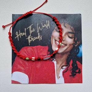 Könnte beinhalten: Rotes Makramee-Armband mit kleinen weißen, schwarzen und grünen Perlen. Das Armband liegt auf einer Karte mit dem Text "Heal The World Threads" und einer lächelnden Person in einer roten Jacke.