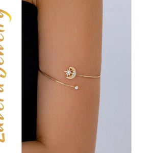 May include: A gold-tone arm cuff featuring a crescent moon and star motif adorned with small clear stones. The open-ended design terminates in a single clear stone at one end. The text Luvera Jewelry is visible on the left side of the image.