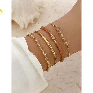 Puede incluir: Un conjunto de cuatro pulseras que combina metal dorado, cuentas transparentes facetadas y cuentas cilíndricas de color marrón claro mate. El set incluye una pulsera de barra dorada lisa, dos pulseras de cuentas con detalles dorados y una de cuentas.