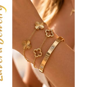 Könnte beinhalten: Ein goldfarbenes Schmuckset bestehend aus zwei zarten Kettenarmbändern mit kleeblattförmigen Anhängern in texturierten und durchbrochenen Designs sowie einem starren goldfarbenen Armreif mit eingelassenen klaren Steinen. Text: Luvera Jewelry.