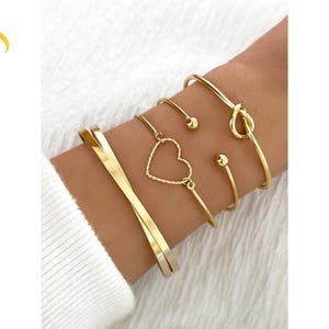 Op de afbeelding: Een verzameling goudkleurige armbanden om een pols. De armbanden omvatten een gedraaide band, een hartvormige bedel en een knoopontwerp. De armbanden worden op een witte achtergrond weergegeven. De tekst "Luvera Jewelry" is zichtbaar.