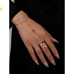 May include: A gold-toned hand chain bracelet featuring a delicate chain that connects a wrist strap to a chevron-shaped ring. The chain is accented with small gold beads and two circular black stones. The text LuveraJewelry is visible on the left.