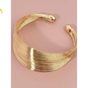 May include: Gold-toned metal cuff bracelet. The open-ended design features multiple parallel wires, creating a layered effect. The bracelet has rounded end caps. The background is a solid, muted pink.