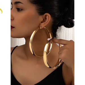 Puede incluir: Grandes pendientes de aro en tono dorado pulido con una superficie lisa y redondeada. Los pendientes presentan un diseño circular clásico con un acabado metálico reflectante. El texto LuveraJewelry es visible a la izquierda en degradado.