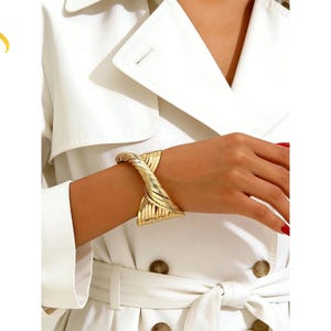 Könnte beinhalten: Ein goldfarbenes Manschettenarmband mit einem gedrehten, mehrsträngigen Design, das in der Mitte zusammenläuft. Das Metall hat eine polierte, reflektierende Oberfläche mit sichtbaren Rillen entlang der Bänder. Der Text LuveraJewelry steht links.