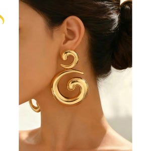 Könnte beinhalten: Goldfarbene Statement-Ohrringe mit einem großen, wirbelnden Design. Die Ohrringe haben eine polierte Oberfläche und eine auffällige Form. Ein auffälliges Accessoire.