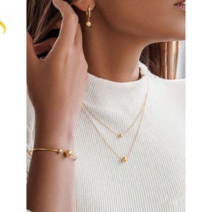 Puede incluir: Un conjunto de joyería dorada que incluye un collar de doble cadena con pequeñas cuentas esféricas, una pulsera abierta a juego con extremos esféricos y un par de pendientes de aro con un dije de cuenta colgante. El texto LuveraJewelry es visible.