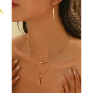 Op de afbeelding: Goudkleurige gelaagde ketting en bijpassende oorbellen. De ketting heeft drie fijne kettingen met staafhangers. De oorbellen zijn eenvoudige staafjes. De sieraden worden door een persoon gedragen.