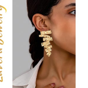 Puede incluir: Un pendiente dorado llamativo que presenta una serie de placas metálicas rectangulares apiladas con un acabado martillado y texturizado. El pendiente tiene un diseño vertical en cascada. El texto Luvera Jewelry es visible a la izquierda.