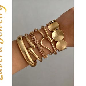 Könnte beinhalten: Ein Set aus goldfarbenen Armbändern mit einer dicken polierten Manschette, einem tropfenförmigen Armreif, einer strukturierten Gliederkette, einem gewellten Armreif, einem geknoteten Armreif und einem Armband mit drei runden Scheiben. Text Luvera Jewelry.