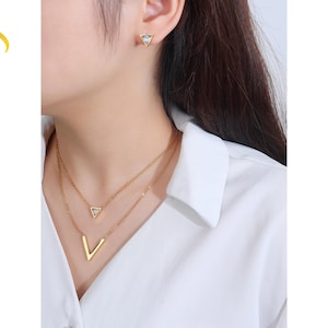Puede incluir: Juego de joyas doradas con un collar en capas y pendientes a juego. El collar tiene un colgante en forma de V y un colgante triangular más pequeño con una piedra transparente. Los pendientes son pequeños tachuelas triangulares.