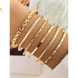 Puede incluir: Un conjunto de seis brazaletes de metal en tono dorado con diversas texturas y diseños, incluyendo un patrón de cuerda trenzada, una banda lisa pulida, un alambre en espiral, una banda geométrica facetada, una barra texturizada y un diseño de rombos.