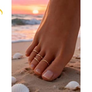 Può includere: Primo piano di un piede su una spiaggia sabbiosa, adornato da tre delicati anelli d'oro per le dita dei piedi. Gli anelli hanno diversi design, tra cui uno stile a corda intrecciata. Lo sfondo presenta un tramonto sull'oceano e il testo "Luvera Jewelry".