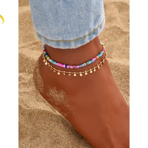 May include: Two anklets on an ankle. The top anklet has colorful beads, and the bottom anklet has small gold-colored circular charms. The background is sand, and the person is wearing blue jeans.