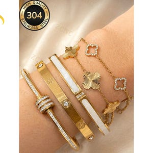 May include: A set of four gold-toned bracelets made of 304 stainless steel. One features a central cylinder with clear stones, another is a plain band with small stones, one has a white inlay, and the last is a chain with clover-shaped charms.