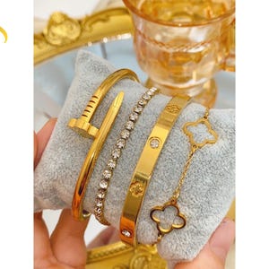May include: A set of four gold-toned bracelets displayed on a light grey velvet cushion. The collection includes a nail-shaped bangle, a crystal-encrusted tennis bracelet, a flat band with clover-shaped cutouts and stones, and a delicate clover chain.