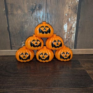 May include: A pyramid stack of six bright orange plastic jack-o'-lanterns. Each pumpkin features a glossy finish with black painted facial details, including triangular eyes, a triangular nose, and a jagged mouth with two teeth on the top row.