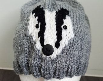 Puede incluir: Un gorro de punto gris con un diseño de cara de tejón. La cara del tejón es blanca con rayas negras, ojos de botón negros y una nariz de botón negra. El sombrero se muestra en una cabeza de maniquí blanca.