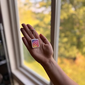 May include: A square keychain featuring the Instagram logo with a white border and a gradient color scheme of purple, pink, orange, and yellow. The keychain is attached to a silver-toned metal split ring by a short chain link connector.