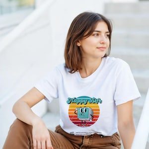 Peut inclure: T-shirt blanc avec un motif rétro d'un écran d'ordinateur de dessin animé et le texte "Happy Life" au-dessus d'un motif de coucher de soleil. Le moniteur a un visage et salue. Le t-shirt est porté par une personne.