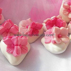 Peut inclure: Une collection de bonbons blancs en forme de cœur, chacun surmonté d'une fleur rose sculptée. Les bonbons sont disposés sur un tissu en maille rose. Le texte www.laconfettata.com est visible au centre de l'image parmi les bonbons.