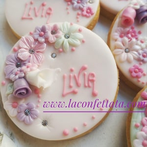 Può includere: Un biscotto rotondo con glassa bianca liscia, decorato con fiori di pasta di zucchero rosa, viola e bianchi, piccole perle argentate commestibili e il nome Livia scritto in glassa rosa. Il testo www.laconfettata.com è stampato in viola.