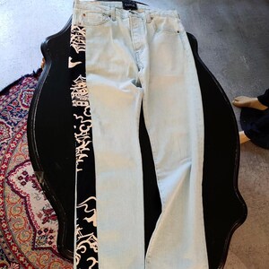 Peut inclure: Jean clair en denim avec un panneau latéral noir orné d'un motif japonais blanc et rouge. Le jean a une conception classique à cinq poches et une fermeture à bouton et fermeture éclair. Le jean est posé à plat sur une surface noire.