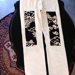 Peut inclure: Jean en denim blanc avec des patchs japonais noirs et blancs au dos. Les patchs présentent un motif de paysage avec des nuages et des bâtiments. Le jean a une conception classique à cinq poches et un patch en cuir marron.