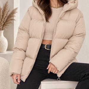 Può includere: Un piumino beige con cappuccio, chiusura frontale con zip e pannelli trapuntati orizzontali. La giacca è indossata sopra un top beige, jeans neri con cintura nera con fibbia dorata e una collana a catena color oro al collo.