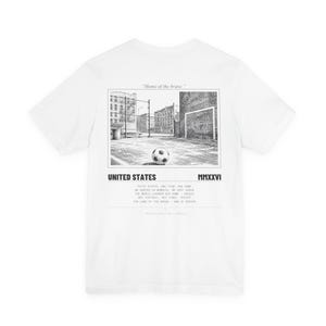 Pode incluir: Camiseta branca com um gráfico a preto e branco de uma bola de futebol num campo com uma baliza. O texto "UNITED STATES" e "MMXXVI" estão abaixo da imagem. O texto adicional inclui "Home of the brave."