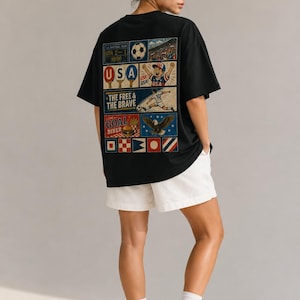 May include: Black t-shirt with a colorful graphic on the back featuring soccer imagery, the text "USA," and the phrase "The Free & The Brave." The shirt is paired with white shorts. The design includes a soccer ball and a stadium.