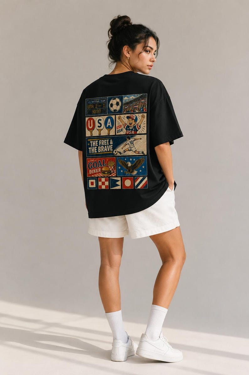 May include: Black t-shirt with a colorful graphic on the back featuring soccer imagery, the text "USA," and the phrase "The Free & The Brave." The shirt is paired with white shorts. The design includes a soccer ball and a stadium.