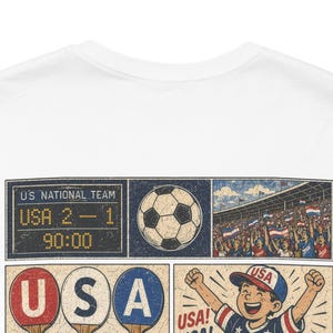 May include: White t-shirt featuring a soccer-themed graphic. The design includes a scoreboard showing a USA score of 2-1, a soccer ball, a crowd waving flags, and a child celebrating with the word "USA!"