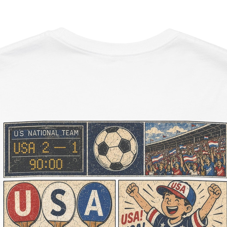 May include: White t-shirt featuring a soccer-themed graphic. The design includes a scoreboard showing a USA score of 2-1, a soccer ball, a crowd waving flags, and a child celebrating with the word "USA!"