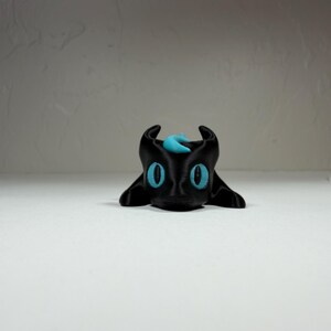 Puede incluir: Una pequeña figura de criatura impresa en 3D de color negro con grandes ojos de color cian brillante y una marca en forma de media luna a juego en la frente. La criatura tiene dos orejas puntiagudas y un acabado mate y suave en el cuerpo.