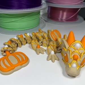 May include: A 3D printed articulated dragon figure in beige with bright orange pumpkin-shaped accents on its tail and head. The dragon features gold-colored trim along its scales, two orange horns, and small white spherical details on its body.