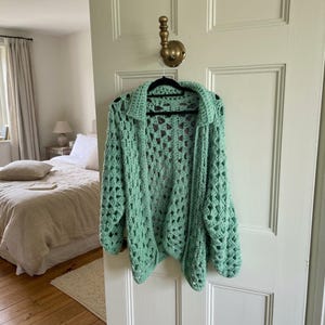 May include: A mint green, open-front cardigan featuring a chunky, loose crochet knit pattern with a diamond-shaped texture. The garment has long sleeves, a folded collar, and a relaxed, oversized fit, hanging from a black hanger on a door hook.