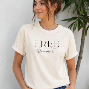 Puede incluir: Camiseta color crema con la palabra "FREE" en negro, una cruz y el texto "Romans 8:1" en letra cursiva. La camiseta tiene las mangas dobladas y se combina con vaqueros azules. Una mujer lleva la camiseta.