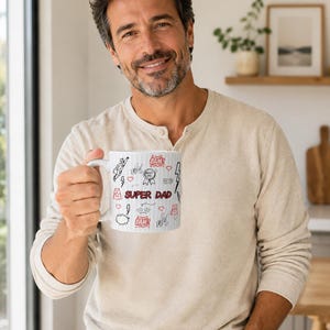 Puede incluir: Taza de cerámica blanca con un diseño "SUPER PAPÁ" en rojo y negro. La taza presenta ilustraciones de dibujos animados de un superhéroe, rayos y corazones. La taza es sostenida por una persona.