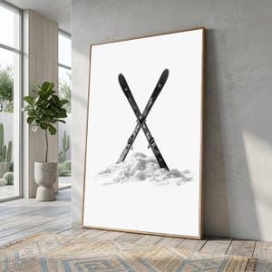 Könnte beinhalten: Ein gerahmter vertikaler Wandkunstdruck mit einer Schwarz-Weiß-Fotografie von zwei in einem Schneehaufen gekreuzten Skiern. Die Skier sind dunkel mit weißen Kreuzsymbolen an den Spitzen und vor einem rein weißen Hintergrund platziert.