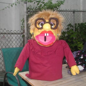 May include: A hand puppet with blonde synthetic hair, wearing large brown plastic glasses and silver stud earrings. The puppet has a yellow face, bright red lips, and a single white tooth, dressed in a long-sleeved maroon shirt with a ruffled collar.