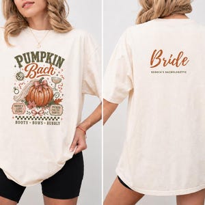 May include: An off-white t-shirt featuring a front graphic with a pumpkin, disco ball, and text reading PUMPKIN BACH, BRIDE'S LAST FLING, PICKED FOR THE PARTY, BOOTS, BOWS, BUBBLY. The back features the text BRIDE and REBECA'S BACHELORETTE.