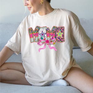 May include: A cream-colored short-sleeve t-shirt featuring the word MAMA in large, multi-patterned letters. The design includes floral, leopard, and polka dot prints with bright stars and a prominent pink ribbon bow graphic centered below the text.
