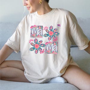 May include: A beige short-sleeved t-shirt featuring a graphic print with the text "Ma" and "Ma" in white letters. The design includes two floral-patterned squares and two large blue and pink flowers, accented with small pink bows and a butterfly.