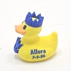 Könnte beinhalten: Eine gelbe Gummiente mit blauer Krone und Akzenten. Die Ente hat einen orangefarbenen Schnabel und schwarze Augen. Das Wort "Allure" und das Datum "7-5-26" sind auf dem Körper der Ente aufgedruckt.