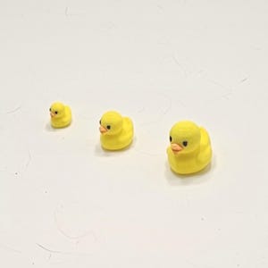 Puede incluir: Tres pequeñas figuras de patos de goma amarillos con ojos negros y picos naranjas. Los patos están dispuestos en fila, disminuyendo de tamaño de derecha a izquierda. El fondo es una superficie lisa de color blanquecino.