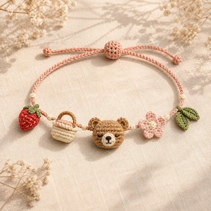 Può includere: Un braccialetto in corda intrecciata rosa con cinque ciondoli all'uncinetto: una fragola rossa, un cestino beige, un muso d'orso marrone con gli occhi chiusi, un fiore rosa chiaro a cinque petali e due foglie verdi, fissato con una perlina rosa.
